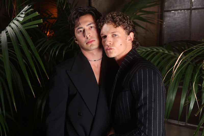 Two handsome male actors in black pose looking directly into the camera, pursing their mouths.