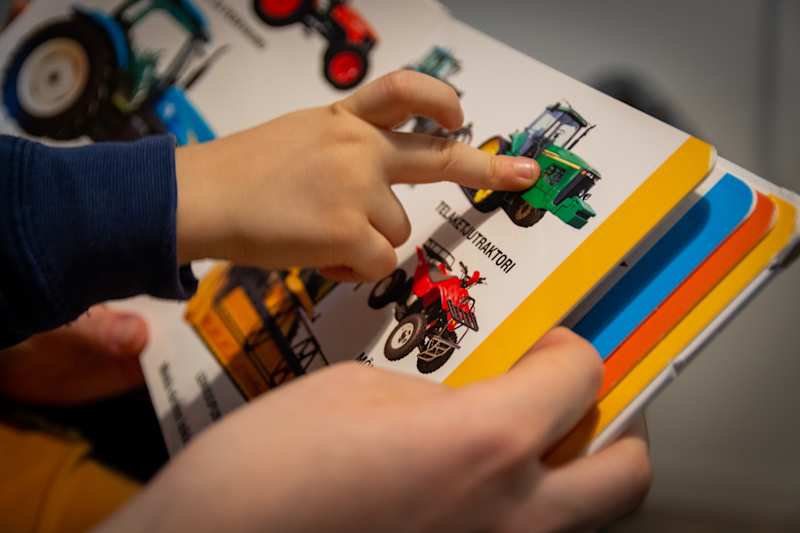 The child's finger points to the tractor on the book page.