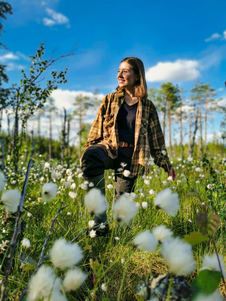 Riikka Karppinen hymyilee kukkien keskellä ruutupaidassa kesäisellä Viiankiaavan suolla.