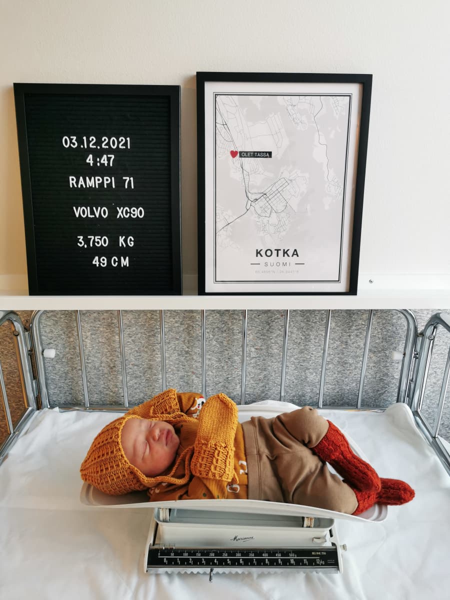 Vauva vaa'alla vaatteet päällä. Vauvan yllä taulut, joissa on merkittynä: 3.12.2021 4:47 Ramppi 71 Volvo XC90 3,750 g 49 cm
