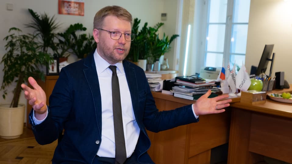 Rybakov puhuu levittäen käsiään. Hänellä on silmälasit, tummansininen puvuntakki, valkoinen kauluspaita ja tumma kravatti. Taustalla näkyy huoneen sisustusta, kukkia. työpöytä.