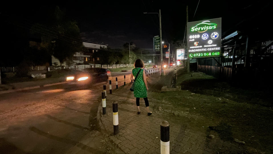 Vihreämekkoinen nainen seisoo selin kameraan öisen kadun varren jalkakäytävällä, autoja tulee vastaan, vieressä vihreä autokorjaamon mainostaulu. 