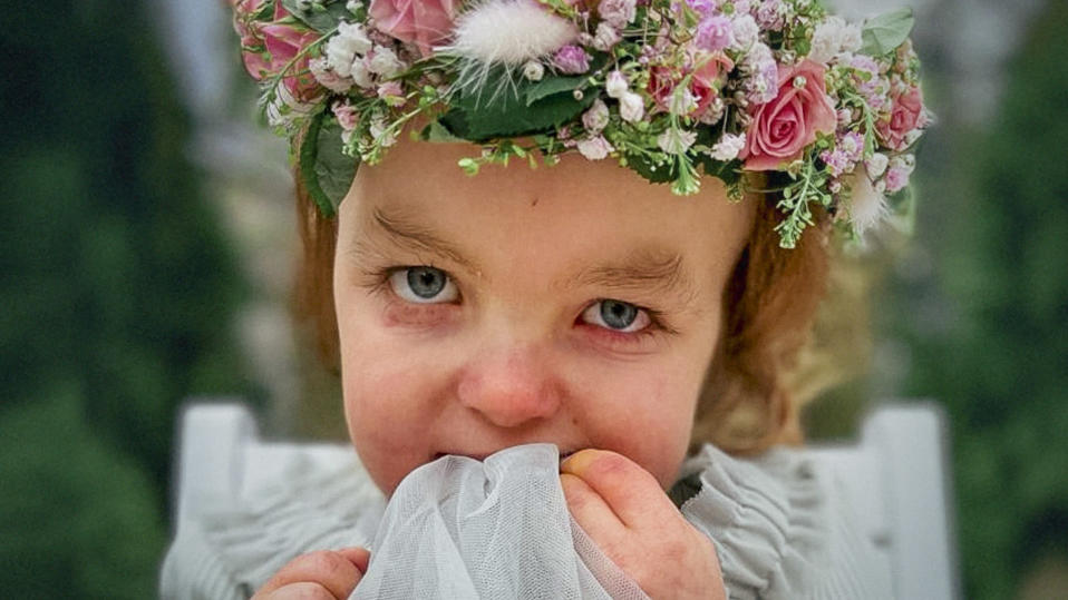 Kuusivuotias Hilda Tolonen katsoo suoraan kameraan kukkaseppele päässään ja hennon vihreän röyhelömekon helma suussaan.