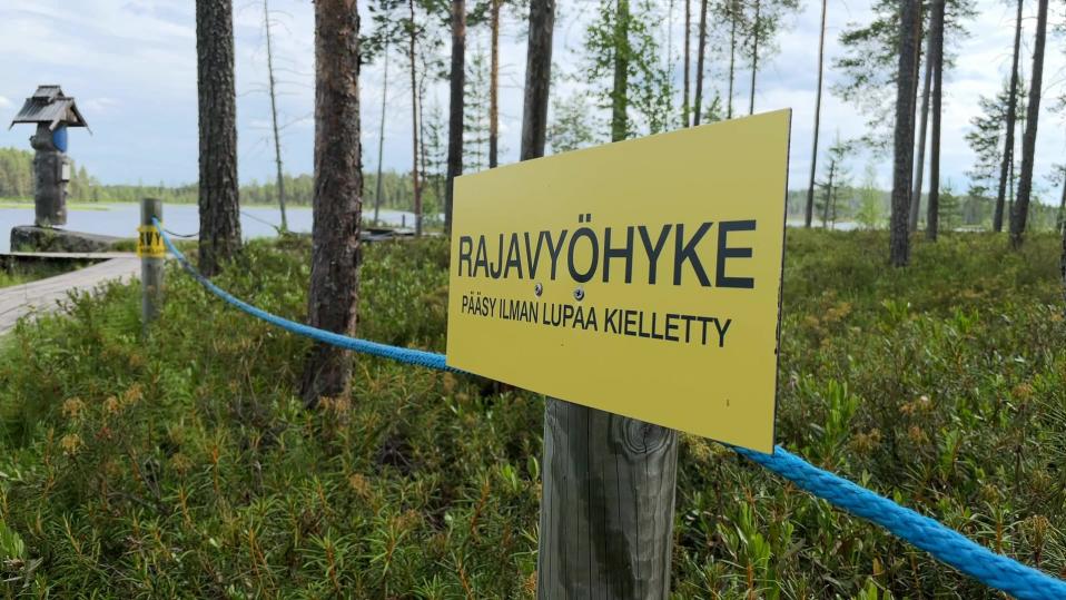 Tolppan kiinnitetty keltainen rajavyöhykekyltti, taustalla itäpisteen puinen merkkipaalu.