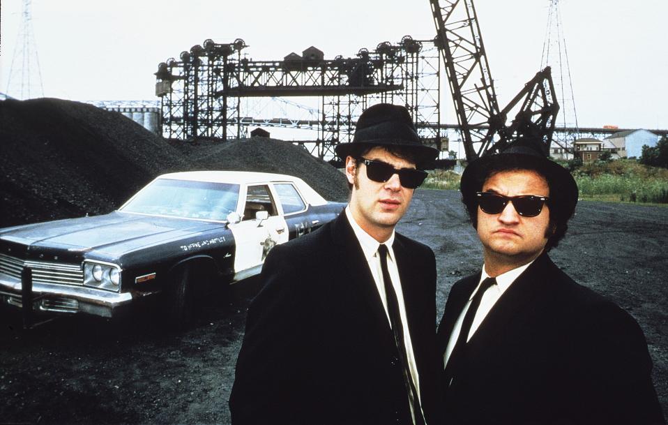 Dan Aykroyd ja John Belushi mustissa puvuissaan. Taustalla vanha poliisiauto ja hiilikasa.