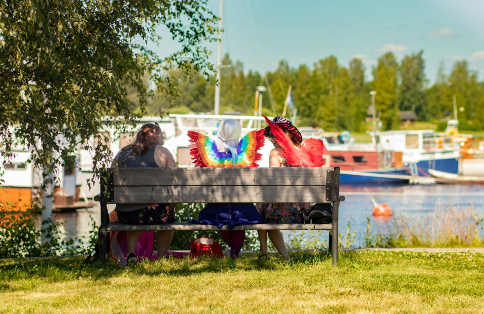 Värikkäisiin asusteisiin pukeutuneita nuoria istumassa järven rannalla penkillä, selkä kameraan päin. Kolmesta nuoresta kahdella on selässään värikkäät siivet.