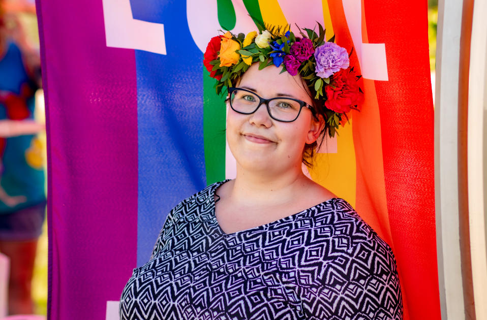 Silmälasipäinen nuori nainen kukkaseppele päässään hymyilemässä kameralle. Taustalla Pride-lippu.