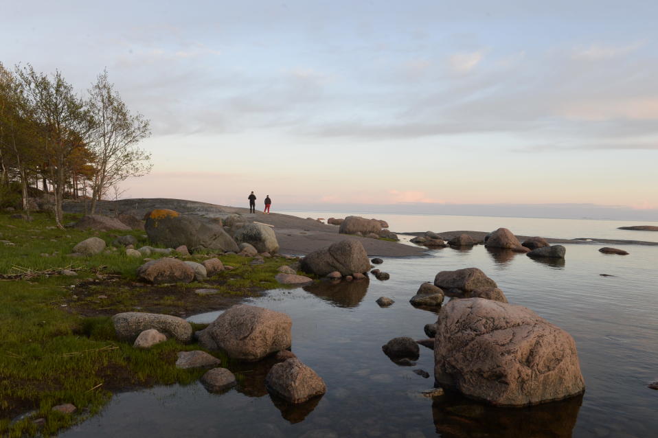 Kaksi ihmistä seisoo selin kivisellä luodolla. Etualalla on rantakiviä. On hieman hämärää ja taivaanranta on punertava.