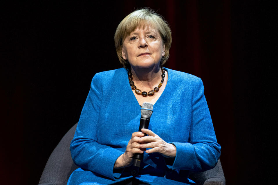 Angela Merkel istuu nojatuolissa spottivalossa ja pitelee mikrofonia. Tausta on musta.