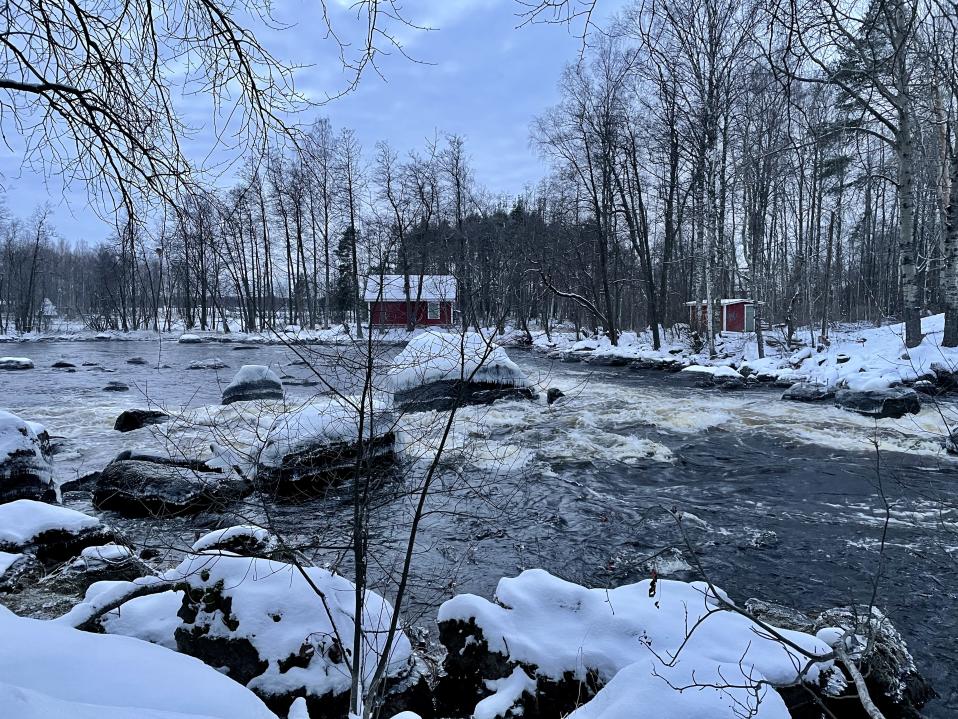 Talvisessa koskimaisemassa punainen mökki ja ulkorakennus.