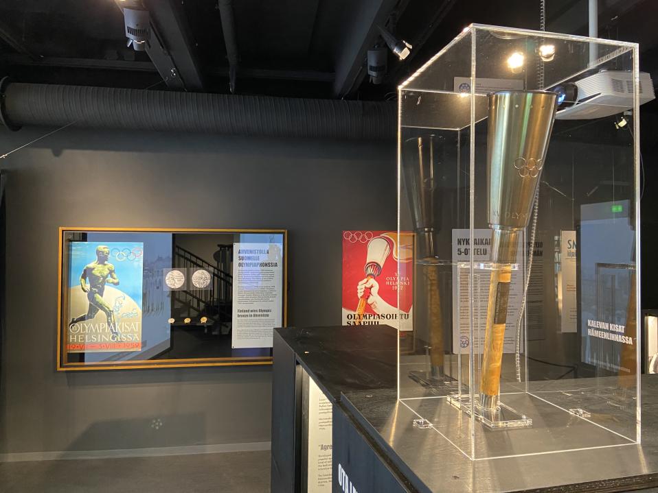 Helsingin olympialaisten Hämeenlinnassa pidettyjen kilpailujen esineistöä Museo Skogsterin näyttelyssä. Kuvassa kopio olympiasoihdusta ja taustalla lasivitriinissä olympiamitalit.