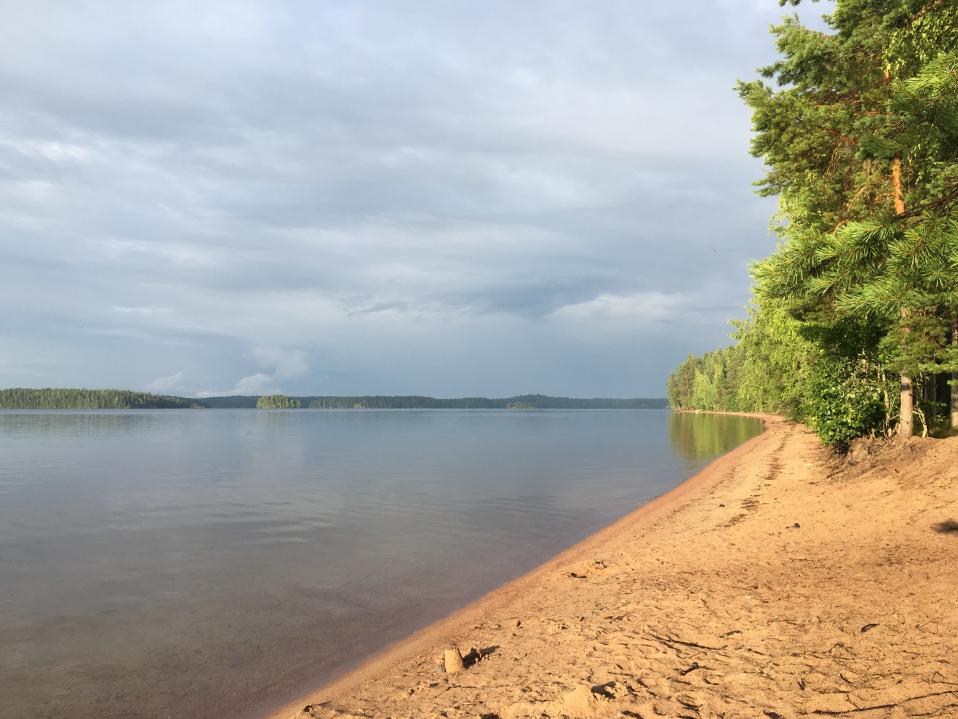 Hiekkarannan rantaviivaa Savitaipaleen Lepänkannon rannassa. Vesi on lähes tyyni, aurinko paistaa mutta taivaalla on tummia pilviä.