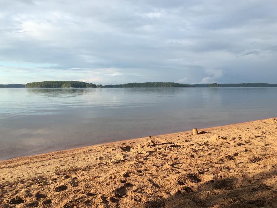 Hiekkarantaa Savitaipaleen Lepänkannossa kuvattuna rannasta Kuolimo-järvelle päin. Vesi on lähes tyyni, aurinko paistaa mutta taivaalla on tummia pilviä.