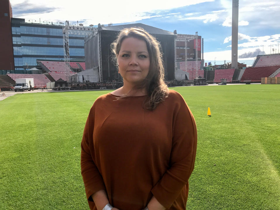 Ruskeahiuksinen nainen seisoo Ratinan stadionin nurmella ja katsoo kameraan. Naisella on yllään ruskea paita, ja taustalla näkyy konserttilava.