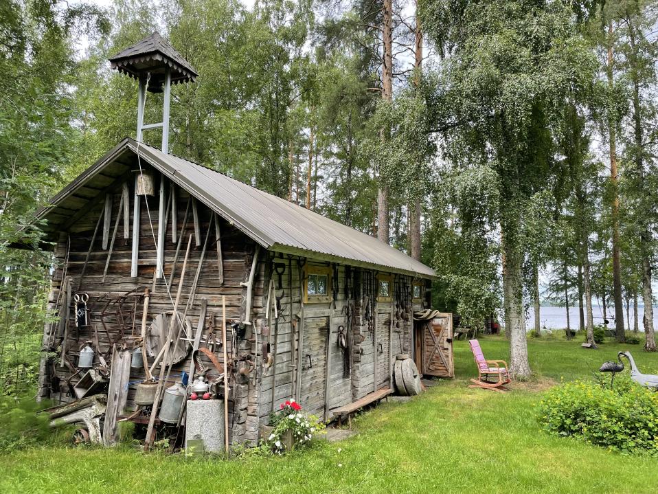 Vanha harmaa aittarakennus järvenrannalla koivikossa, aitan seinillä on paljon vanhoja puisia työkaluja.