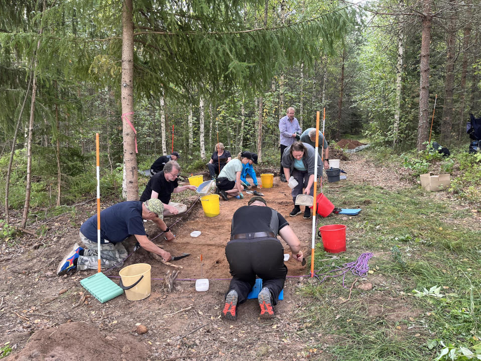 Arkeologiseen kaivaukseen osallistuvia ihmisiä metsän reunassa sijaitsevalla kaivauspaikalla.