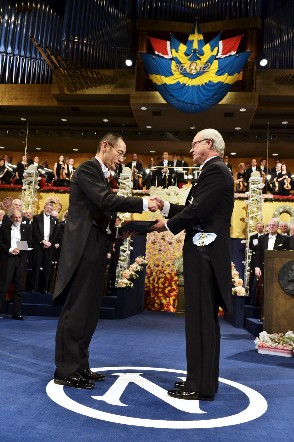 Ruotsin kuningas Kaarle XVI Kustaa ojentaa Shinya Yamanakalle Nobelin palkinnon vasemmalla kädellä. Miehet kättelevät samaan aikaan oikeilla käsillään. Taustalla näkyy juhlapukuihin pukeutuneita ihmisiä.