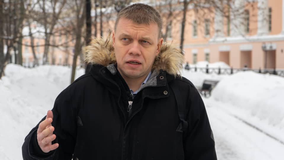 Stupin puhuu talvisessa puistossa. Hänellä on tumma takki, jossa on turkiskaulus. Taustalla näkyy Moskovan duuman eli kaupunginvaltuuston rakennus.