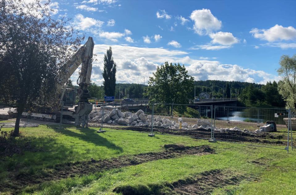 Työmaa-aita, betoninpaloja maassa, kaivinkone. Taustalla Kymijoki ja silta.