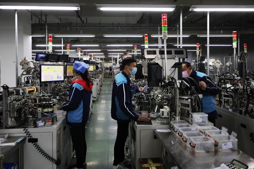 Kuvassa kolme työntekijää kiinalaisella komponenttitehtaalla. Työntekijät seisovat työpisteiden ääressä. 