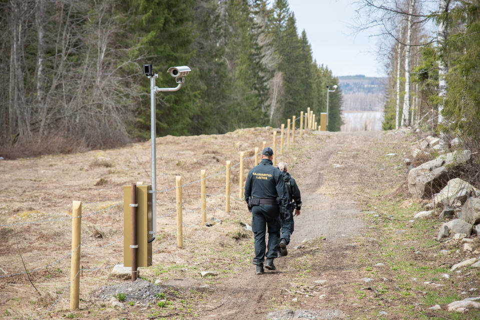 Kaksi rajavartijaa kävelee raja-aukolla piikkilanka-aidan ja valvontakameratolpan vieressä.