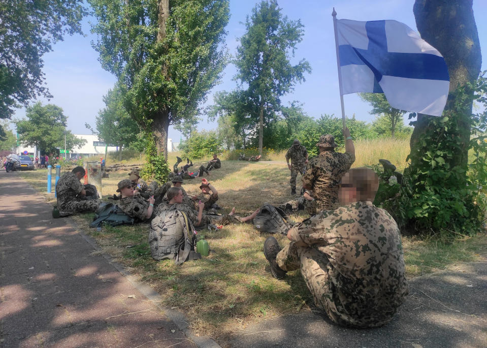 Suomalaisia reserviläisiä istuu puun alla varjossa marssitapahtuman tauolla Hollannissa. Yhdellä kädessä Suomen lippu, joka liehu tuulessa.