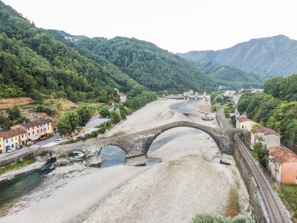 En nästan helt uttorkad floddal i ett italienskt landskap med grönskande kullar och gulnande åkerplättar. Över floden går en bro med flera valv och längs den ena stranden går en enspårig järnväg.