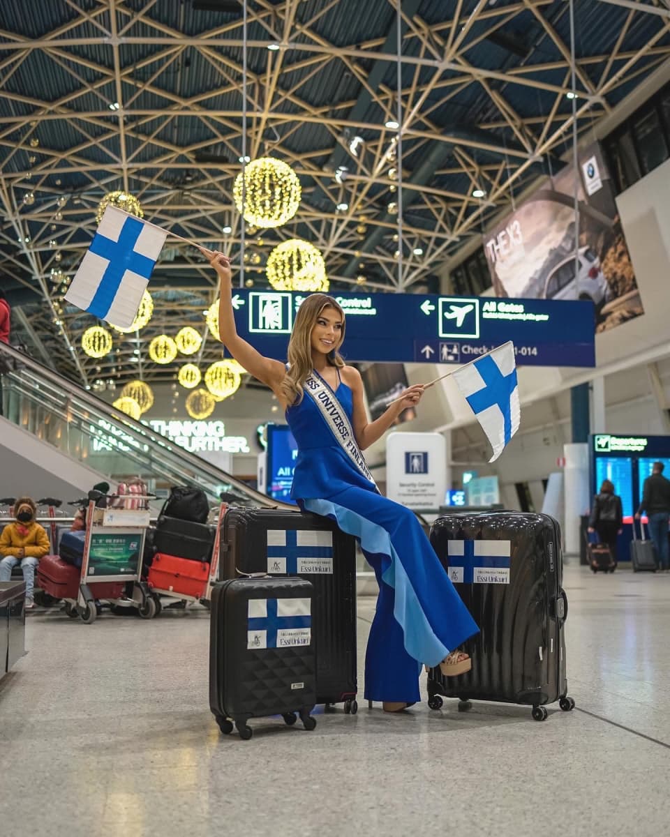 Essi Unkuri poseeraa lentokentällä kameralle sinisessä iltapuvussa, istuu matkalaukkujen päällä ja pitelee Suomen lippua.