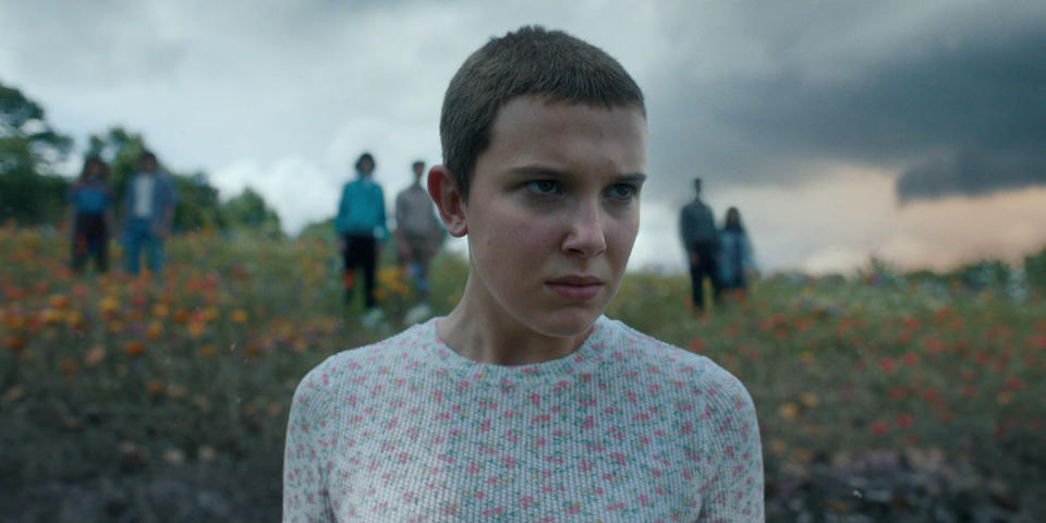 Eleven (Millie Bobby Brown) niityllä, taaempana näkyy kuusi muuta henkilöä.