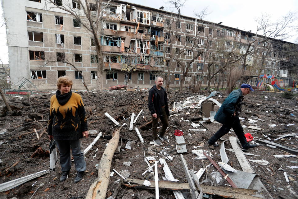 Kolme henkilöä kävelee pommituksissa tuhoutuneen kerrostalon pihalla.