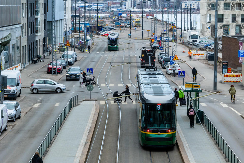 Laajassa kaupunkimaisemassa näkyy raitiovaunuja, lastenvaunujen kanssa suojatietä ylittäviä henkilöitä, autoja ja pyöräilijöitä liikkeessä.