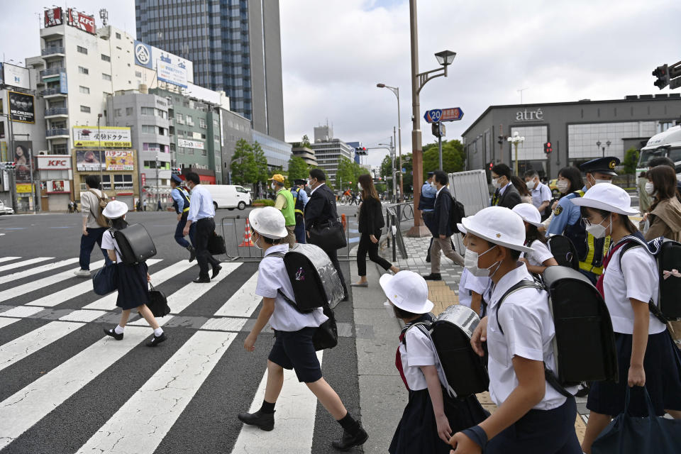 Koululaiset ylittävät tien koulupuvuissa: valkoiset t-paidat, mustat polvipituiset housut, musta reppu ja valkoinen hattu.