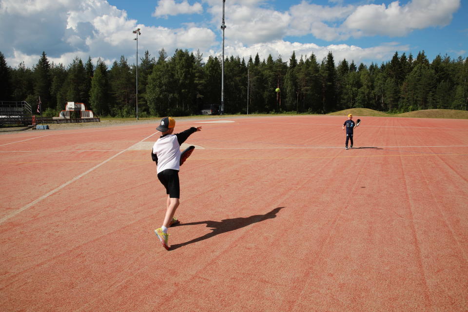 Kaksi lasta heittelee pesäpalloa punaruskealla kentällä, taustalla metsää. 