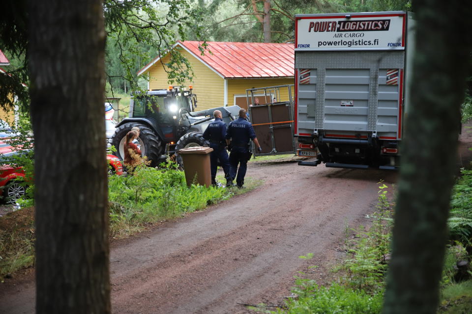 Traktori nostaa suurta laatikkoa kuorma-autoon maatalon pihalla. Kaksi poliisia katsoo nostamista.