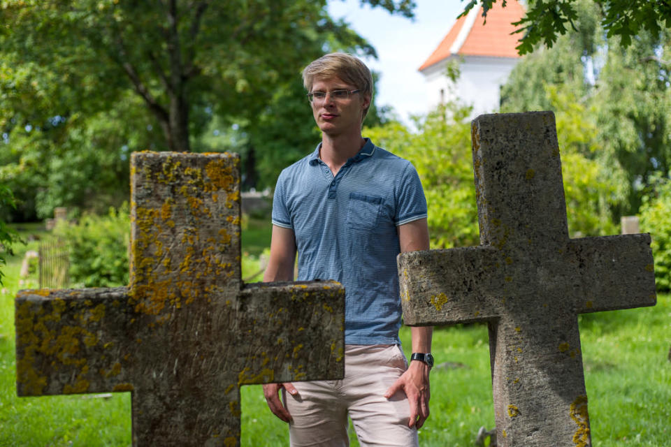 Sinipaitainen vaaleatukkainen ja silmälasipäinen mies seisoo vanhalla hautausmaalla kahden kiviristin välissä.
