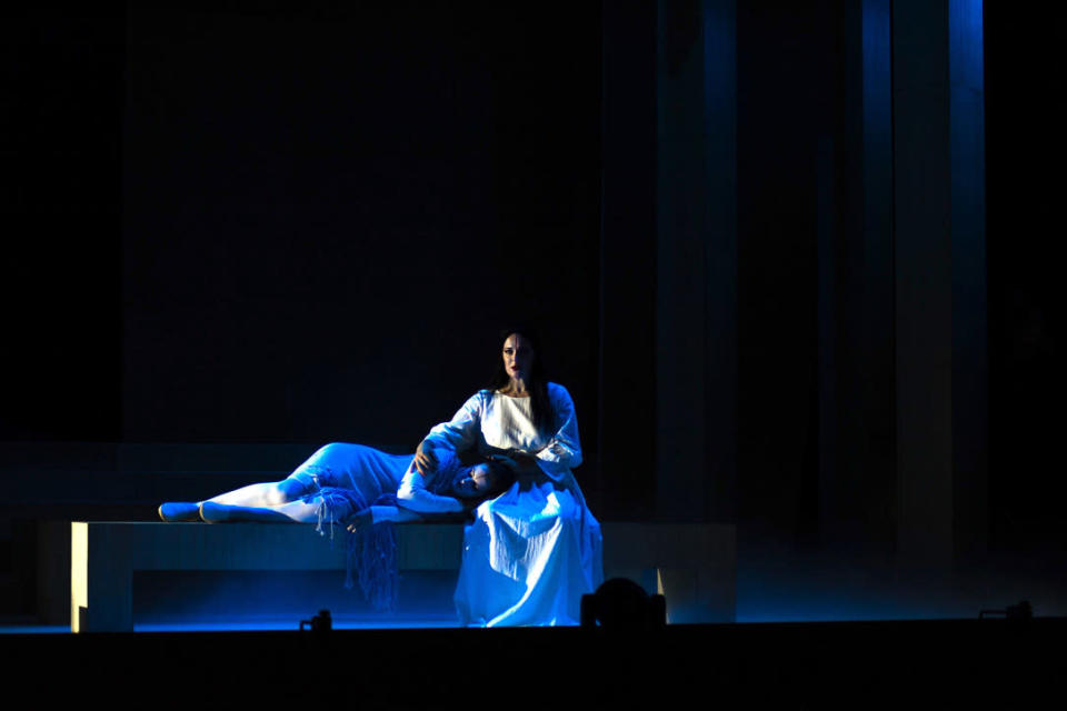 Tummatukkainen nainen istuu ja laulaa pimeällä näyttämöllä toinen nainen makaa penkillä pää laulavan naisen sylissä.
