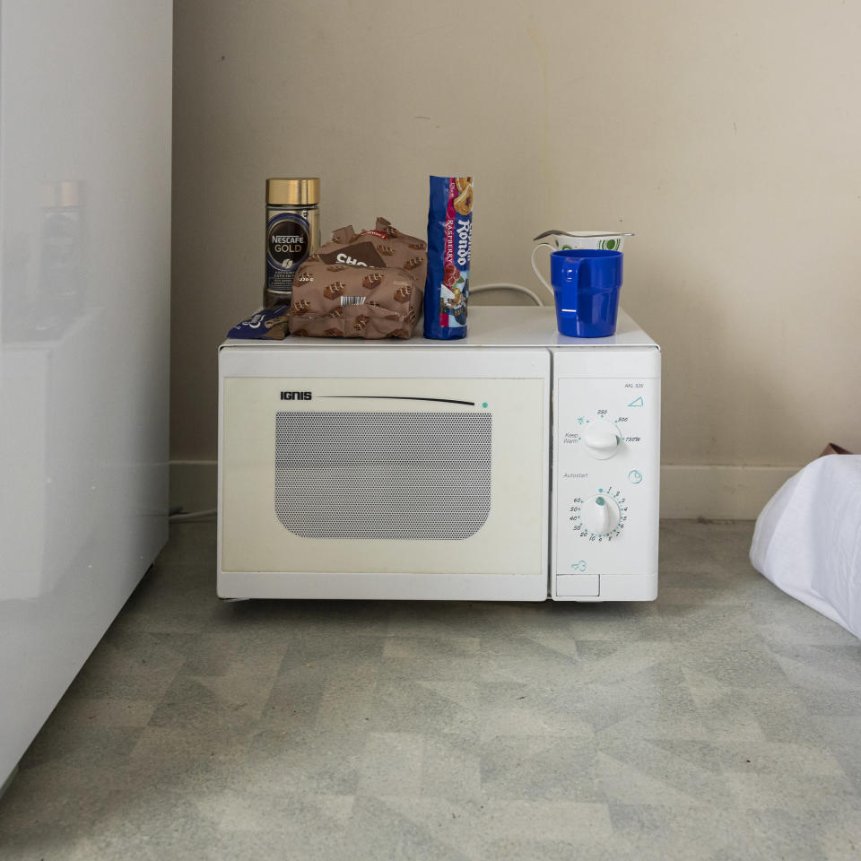 The floor of the refugee women’s room has a microwave with biscuit packs, a jar of instant coffee powder and two mugs.  Refrigerators next to the microwave and a mattress on the floor. 